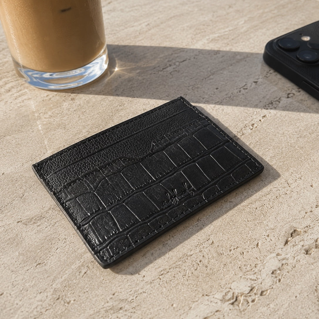 Black textured cardholder on a stone surface with a glass of iced coffee and phone in the background.