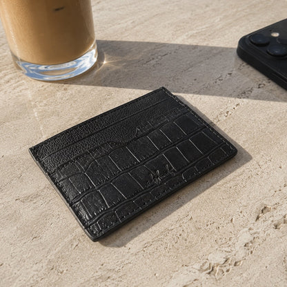 Black textured cardholder on a stone surface with a glass of iced coffee and phone in the background.